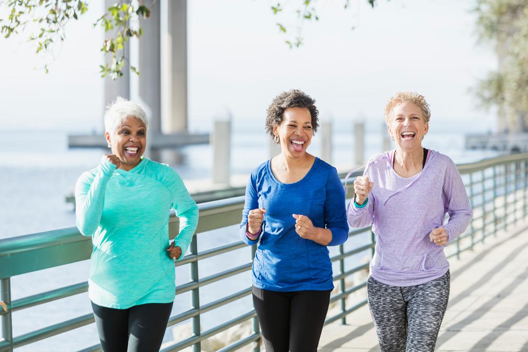 A group of three multi-ethnic mature and senior women in their 50s and 60s exercising, jogging or power walking on a city waterfront side by side. They are having a good time, laughing.
